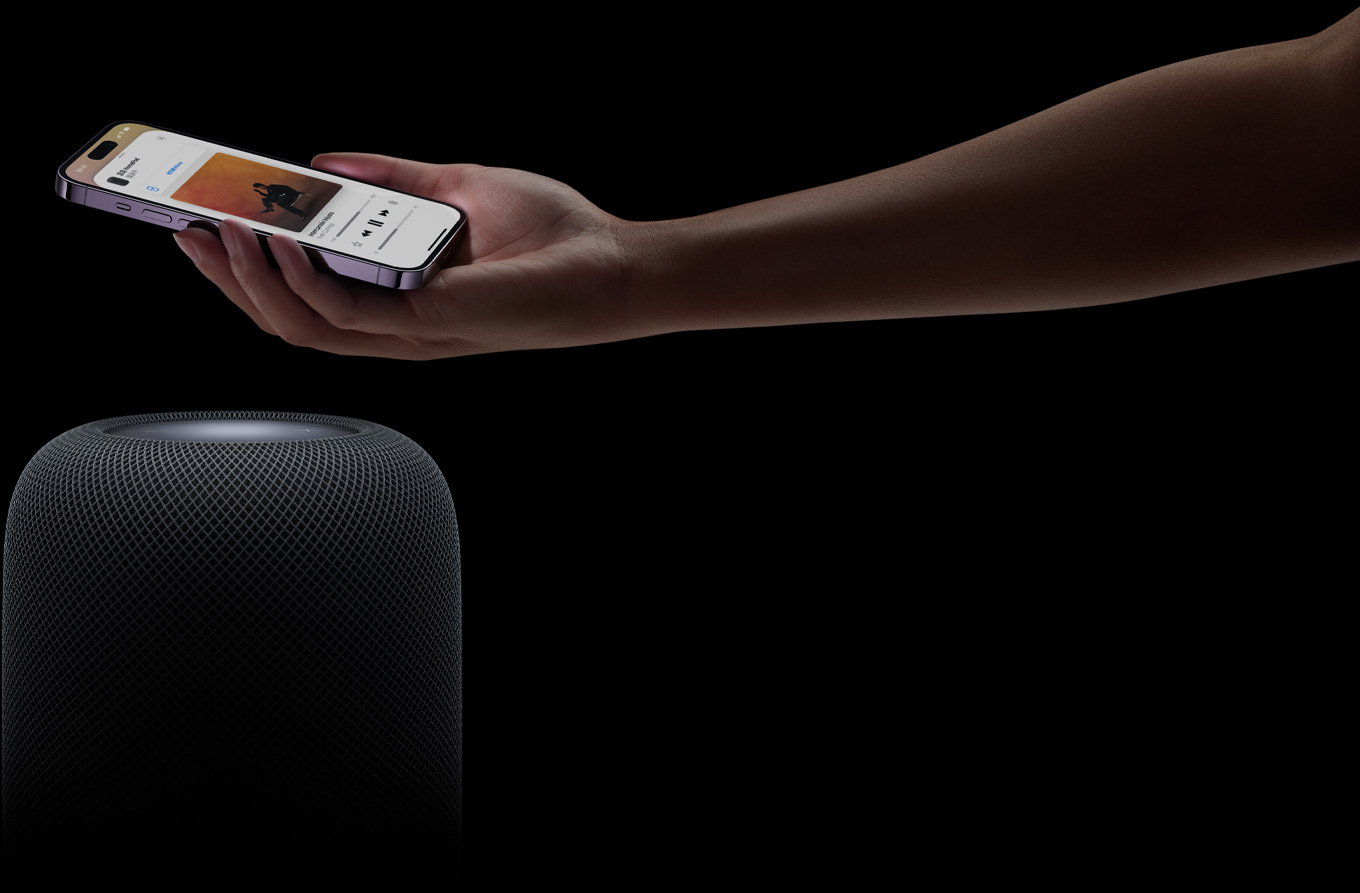 從右側(cè)進(jìn)入頁面的一只手，拿著 iPhone 舉在一個 HomePod 揚(yáng)聲器上方