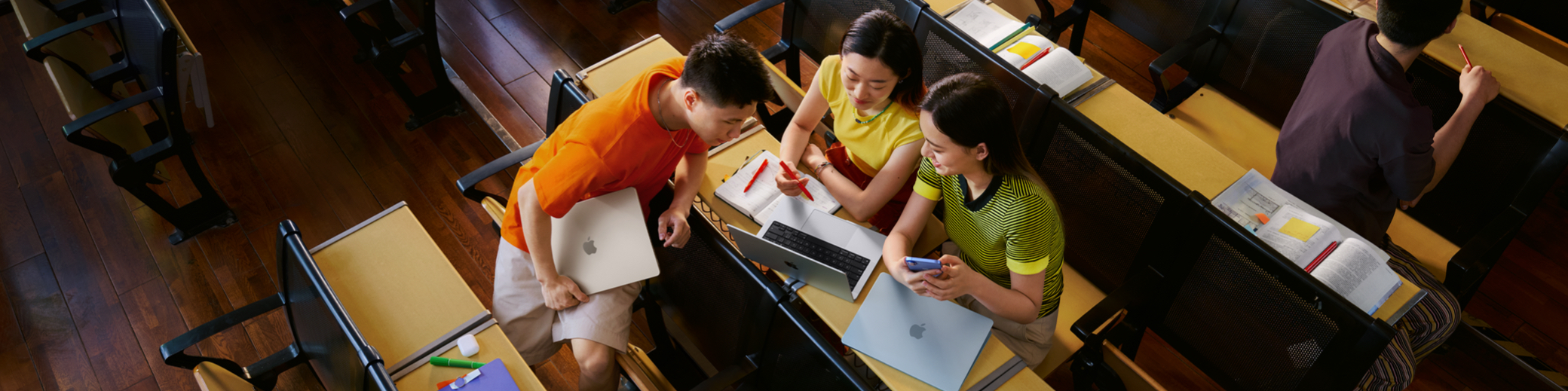 大學(xué)教室的俯拍照片，展現(xiàn)三個(gè)學(xué)生人手一臺(tái) MacBook，其中一位女生的 MacBook 打開(kāi)放在課桌上，他們正圍著屏幕進(jìn)行討論。