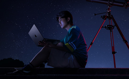 照片展示星空下，一位男生坐在天文望遠(yuǎn)鏡前用著 MacBook Air。