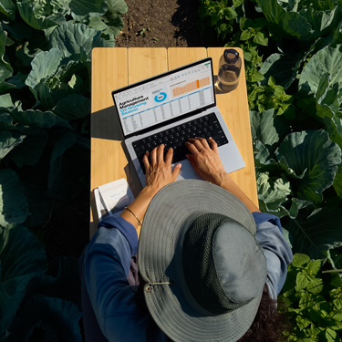 俯瞰圖展示一位戴寬檐帽的女子坐在花園的桌子旁，正在使用 MacBook 制作商務(wù)演示文稿。