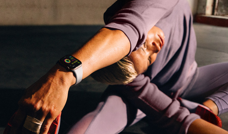 一位女士戴著 Apple Watch Series 10 坐在地板上，手臂向腿部進(jìn)行拉伸，屏幕上顯示體能訓(xùn)練 app 中的整理放松、健身記錄和健身圓環(huán)。