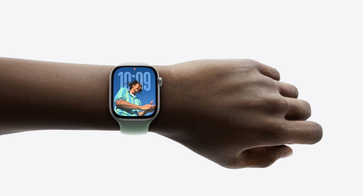 特寫畫面顯示一只手腕戴著配有橡膠表帶的銀色 Apple Watch，表盤展示全新 Liquid Glass 設(shè)計(jì)的時間 app。