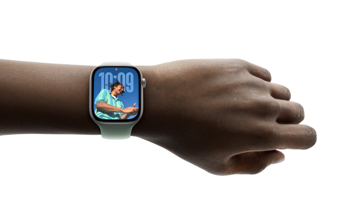 特寫畫面顯示一只手腕戴著配有橡膠表帶的銀色 Apple Watch，表盤展示全新 Liquid Glass 設(shè)計(jì)的時間 app。